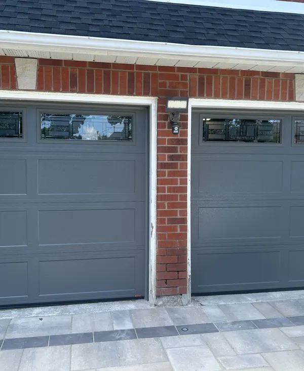 Two gray insulated garage doors with window tops on a brick home