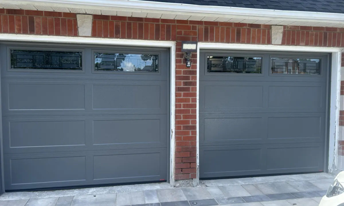 Two gray insulated garage doors with window tops on a brick home